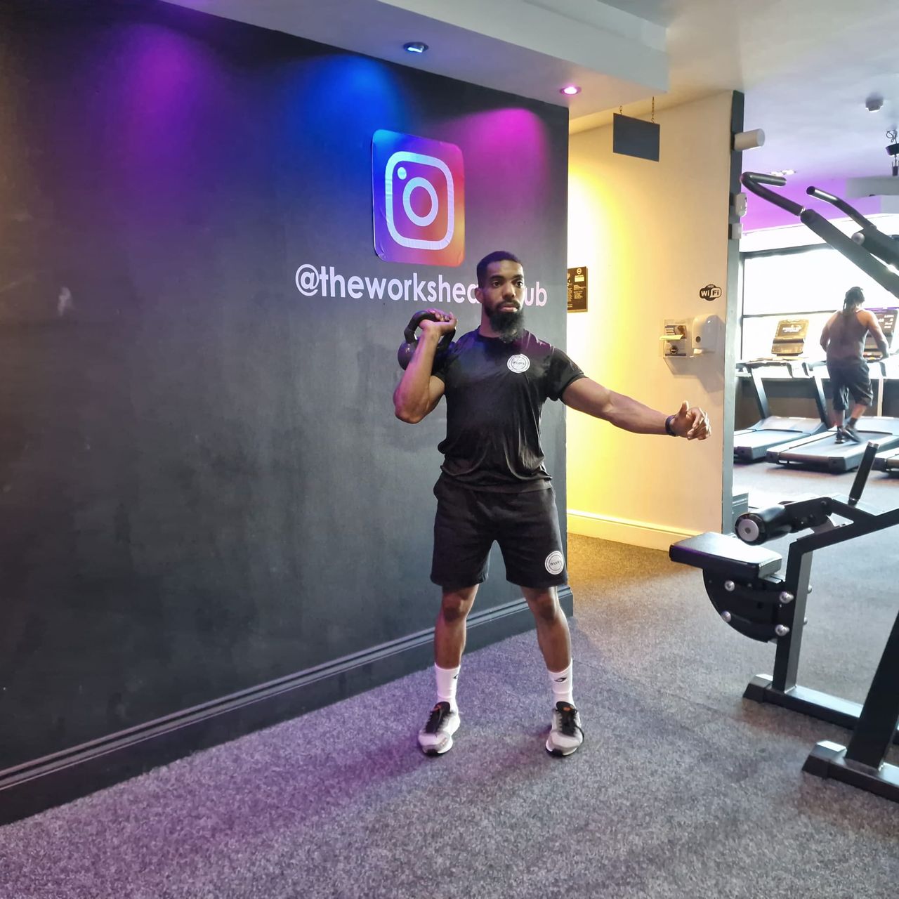 Renel Scarlett performs a kettlebell snatch in The Works Health Club gym. He is standing, feet hip-width apart, with one arm held out to the side and the other bent at the elbow, holding a kettlebell close to his shoulder. Behind him we see a person on a treadmill.
