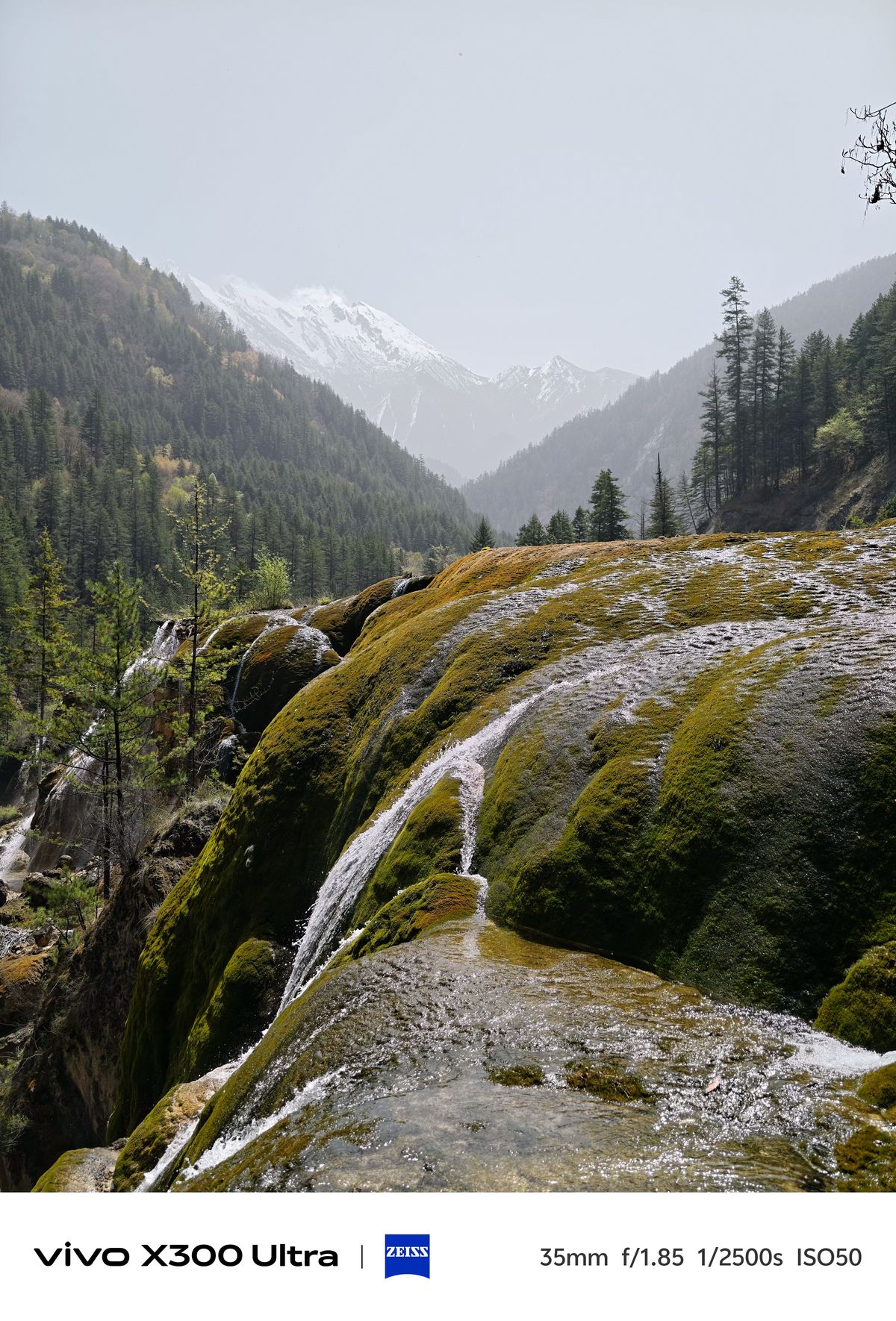 A cascading waterfall flowing over smooth, moss-covered mineral formations against a hazy backdrop of snowy peaks.