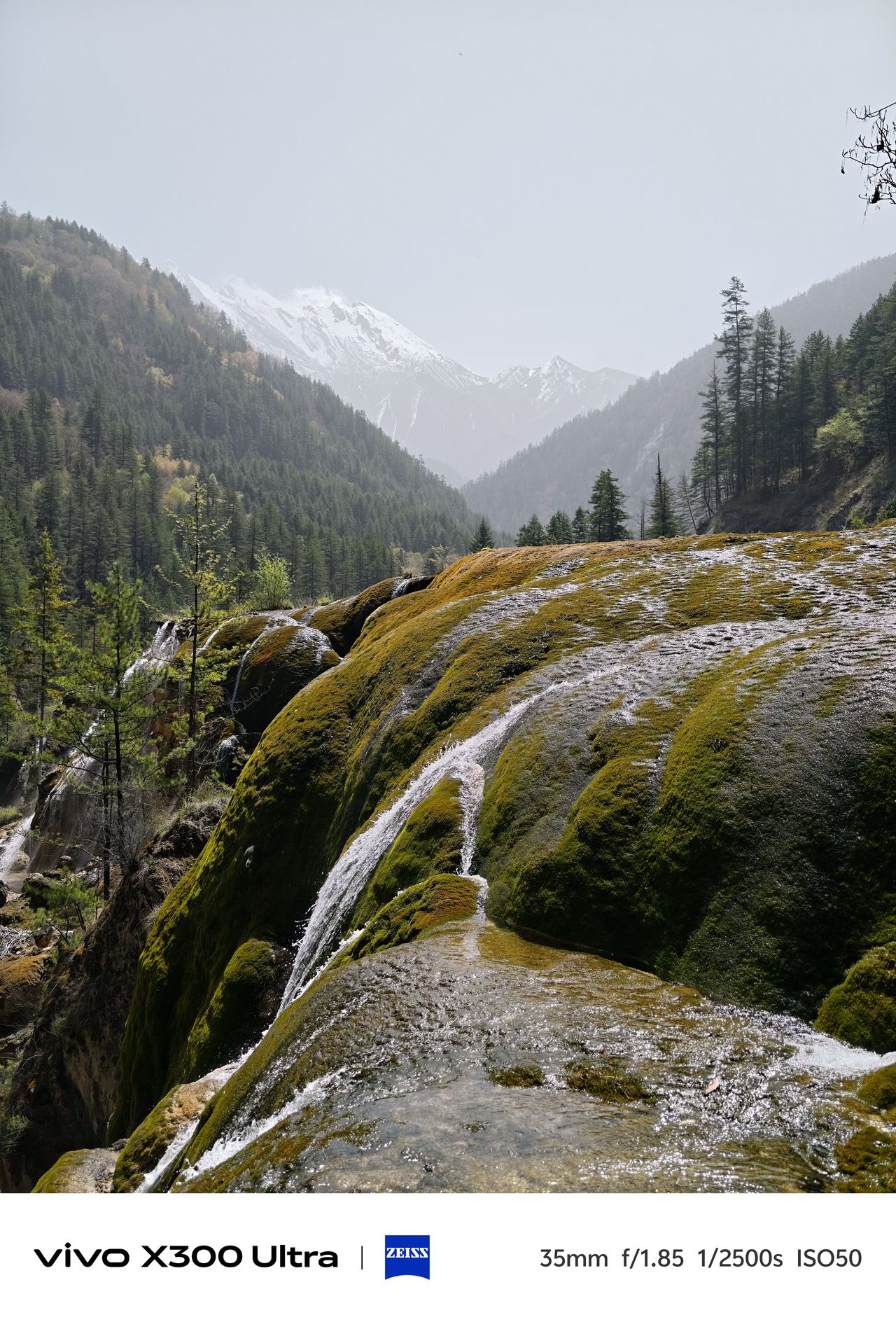 A cascading waterfall flowing over smooth, moss-covered mineral formations against a hazy backdrop of snowy peaks.