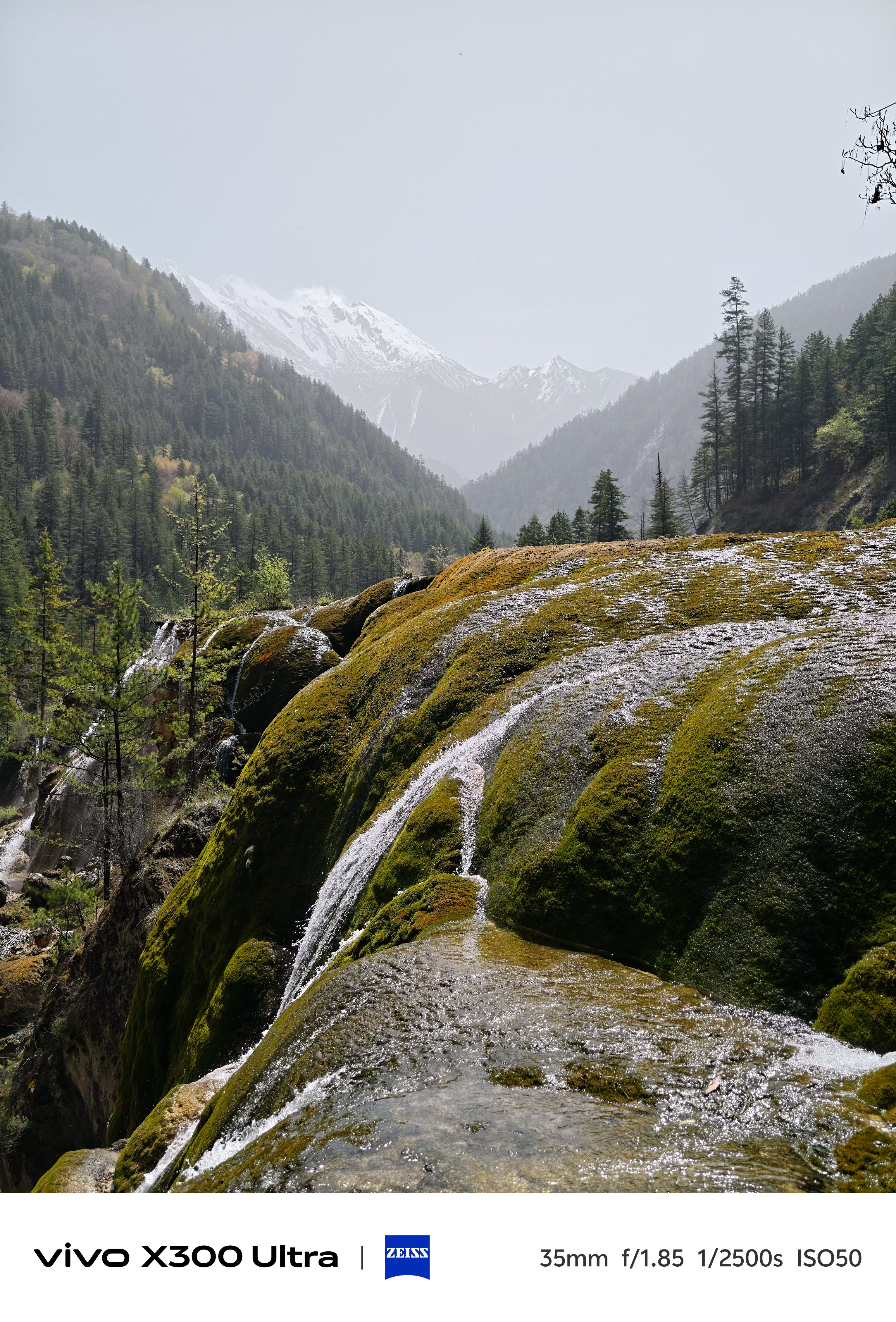 A cascading waterfall flowing over smooth, moss-covered mineral formations against a hazy backdrop of snowy peaks.