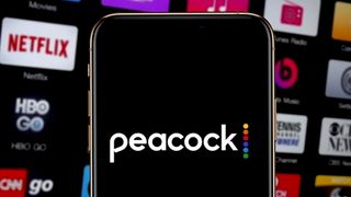Smartphone showing Peacock TV logo in front of blurred out TV screen with other streaming app icons like Netflix, etc