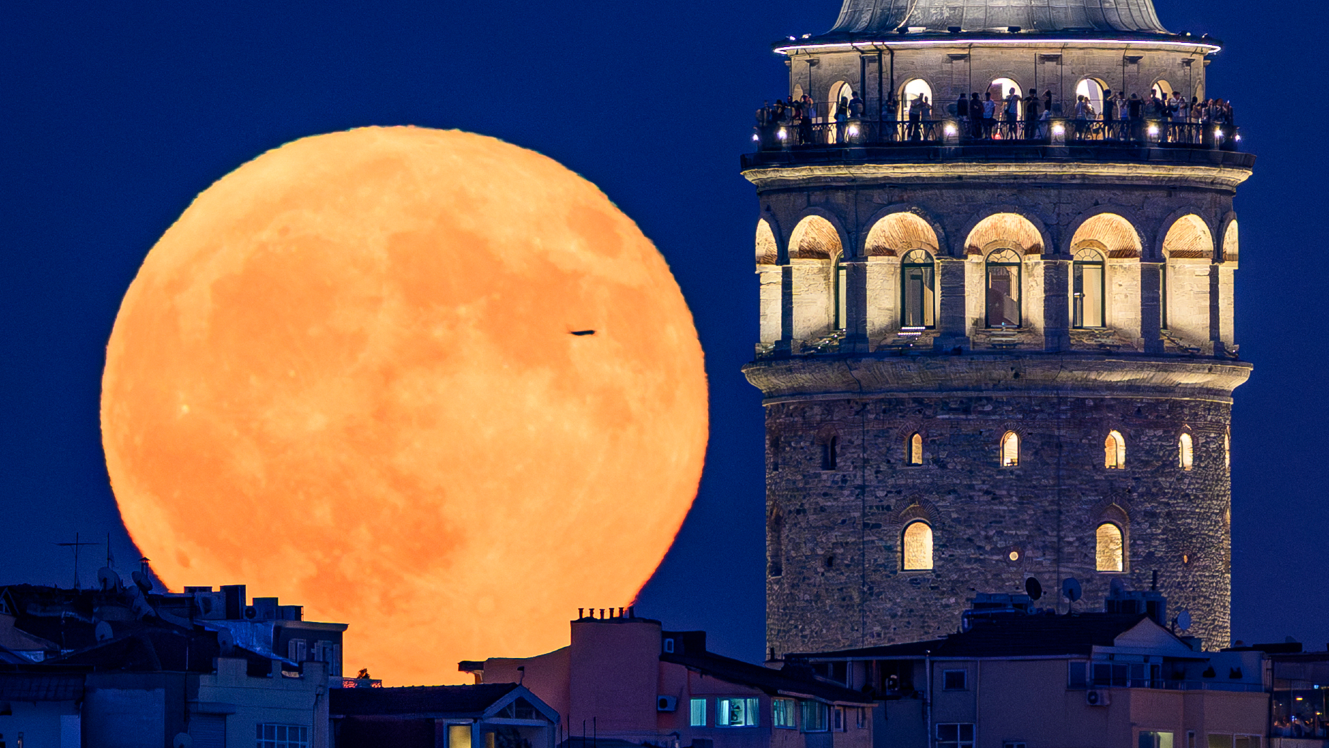 An orange full moon is pictured rising above city rooftops against a dark sky. A castle-like tower dominates the right hand side of the picture, lit by warm light.