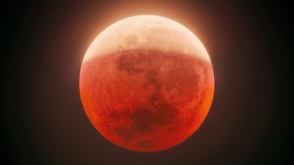 A close up of a bright orange blood moon in the night sky, the top half more yellow than orange