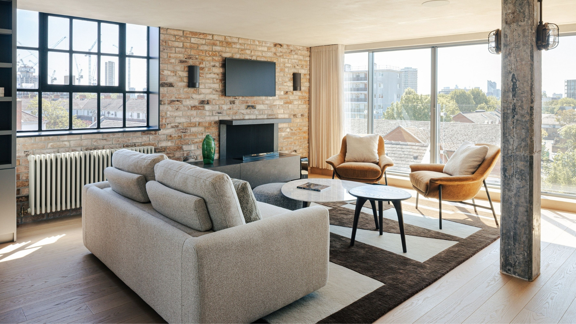 Loft living room with exposed brick wall, grey sofa, mustard armchairs, and large city-view windows.