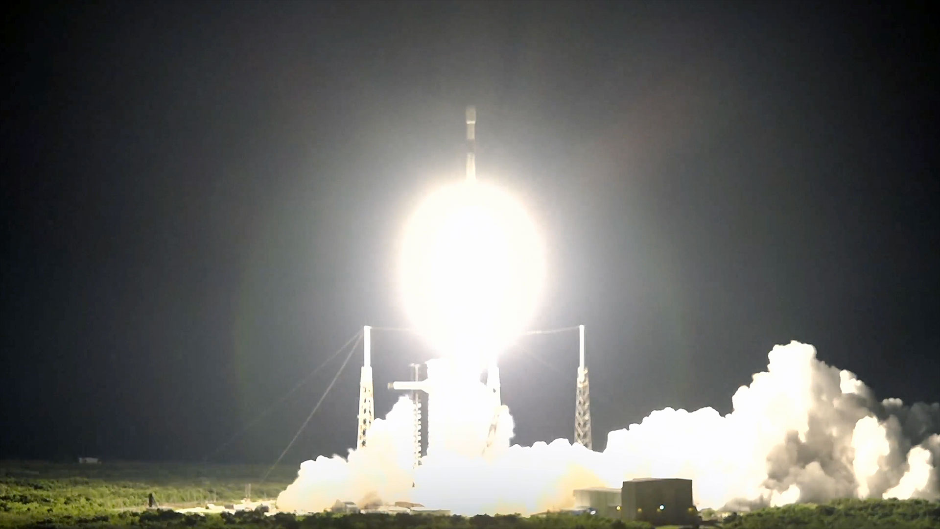 a white and black rocket launches into the dark of night, its bright white thrust lighting up its launch pad and surrounding area.