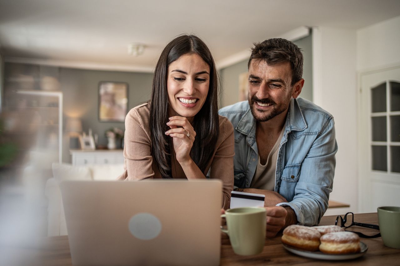 A smiling couple works on their computer in the kitchen, looking at 401(k) balance.