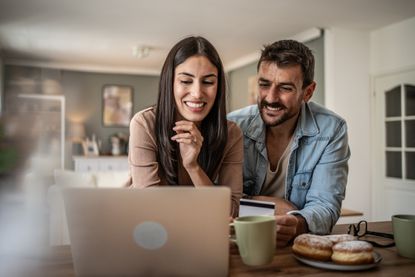 A smiling couple works on their computer in the kitchen, looking at 401(k) balance.