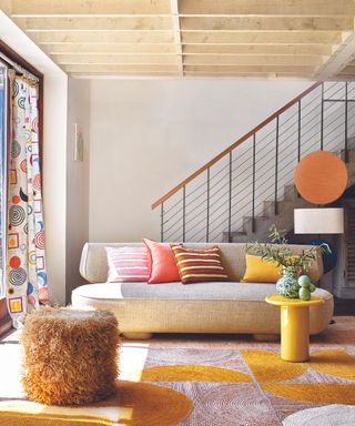 A brightly lit living room with a white curved couch, bright pops of color and a stairway in the back