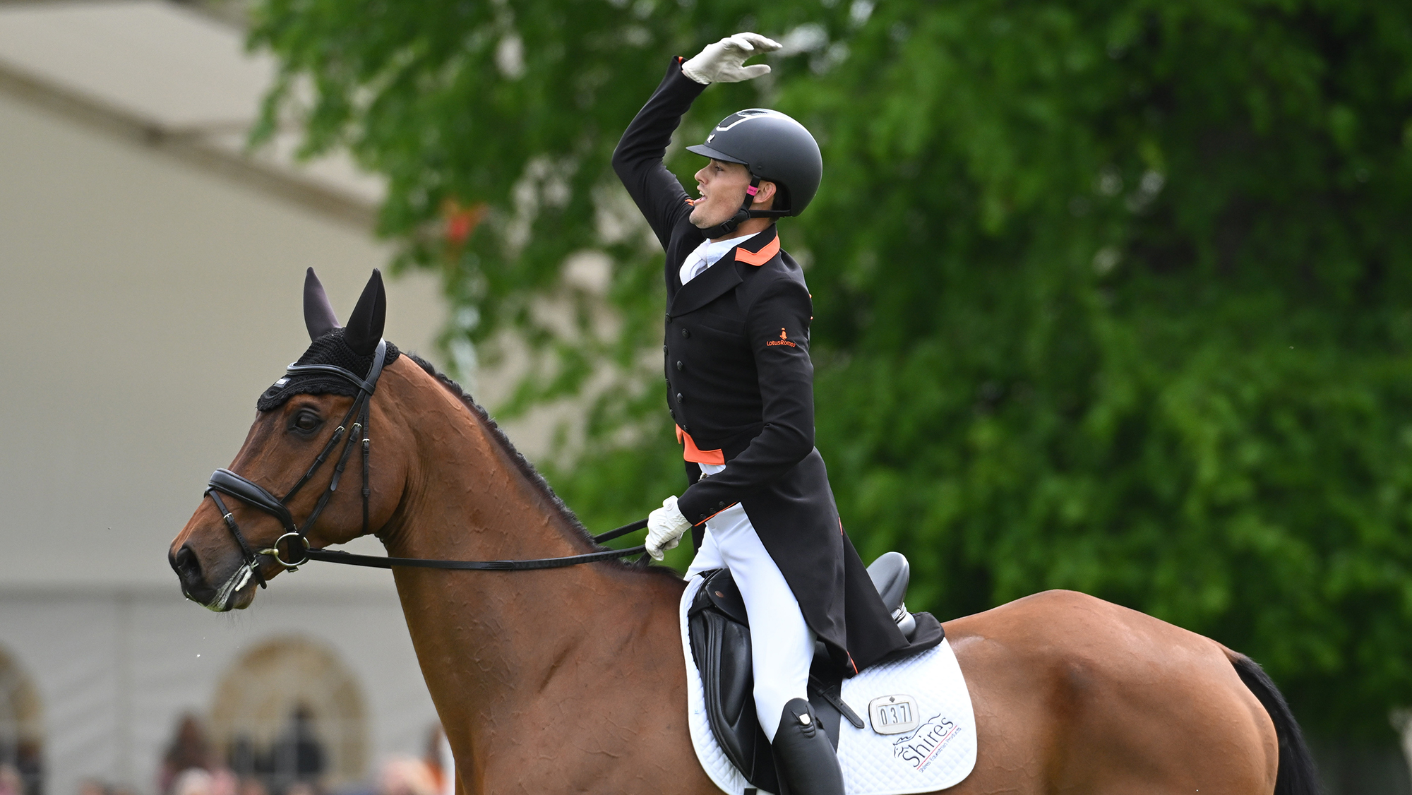 Tom Woodward, a rider in a black tailcoat with orange trim, raises a hand in triumph after a dressage test on Low Moor Lucky, a bay horse, at Badminton Horse Trials 2025.