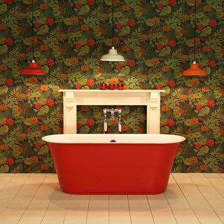 Red freestanding bath on top of light wooden flooring, with two red and one white pendant lights above it, in front of a green and red floral wallpaper