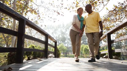 An older couple smile and hold hands as they walk across a wooden bridge.