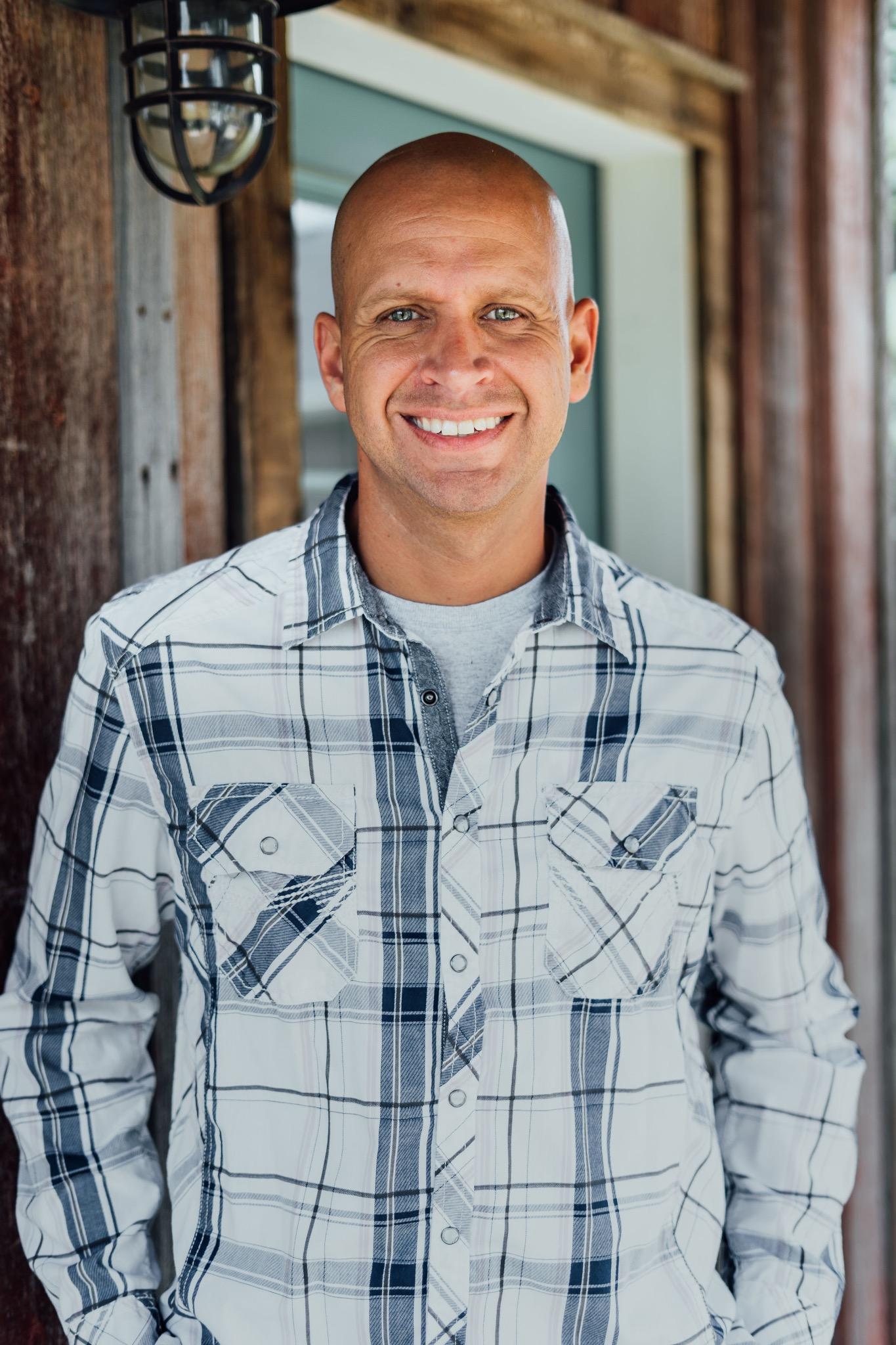 Dan is a man wearing a collared checked shirt, standing on a wooden porch