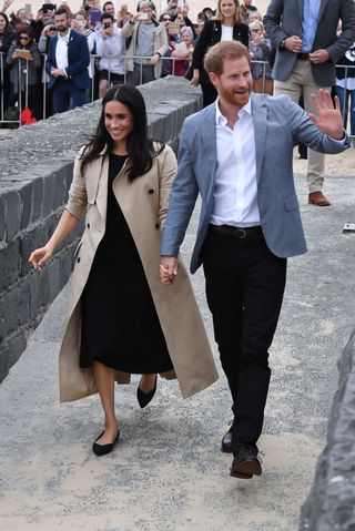 Meghan Markle wearing a tan coat and black dress with black Rothy's flats holding hands with Prince Harry walking past fans and waving