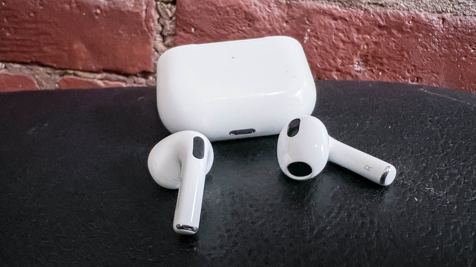 Apple AirPods 3 with charging on a black leather surface against a brick wall backgound