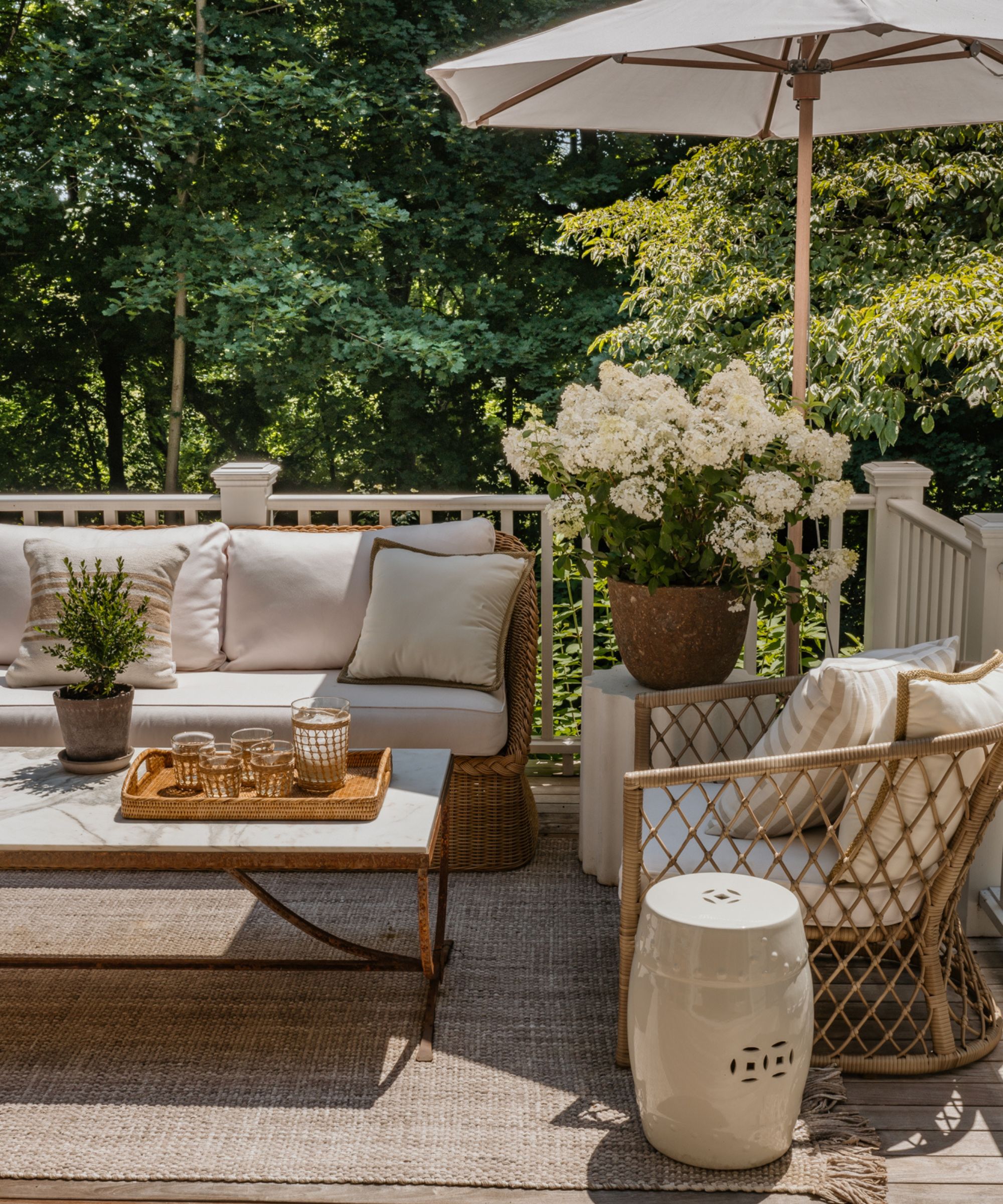Porch with a jute rug, a rattan sofa and armchair, a marble and iron coffee table, a cream parasol, and a large potted plant