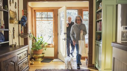 An older couple smile as they come through the front door of their home after shopping.