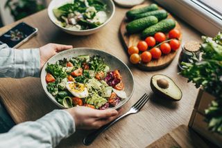 A pair of hands holding a large bowl of delicious salad with leafy greens, tomatoes, avocado and egg, surrounded by a kitchen surface with items of salad on top.