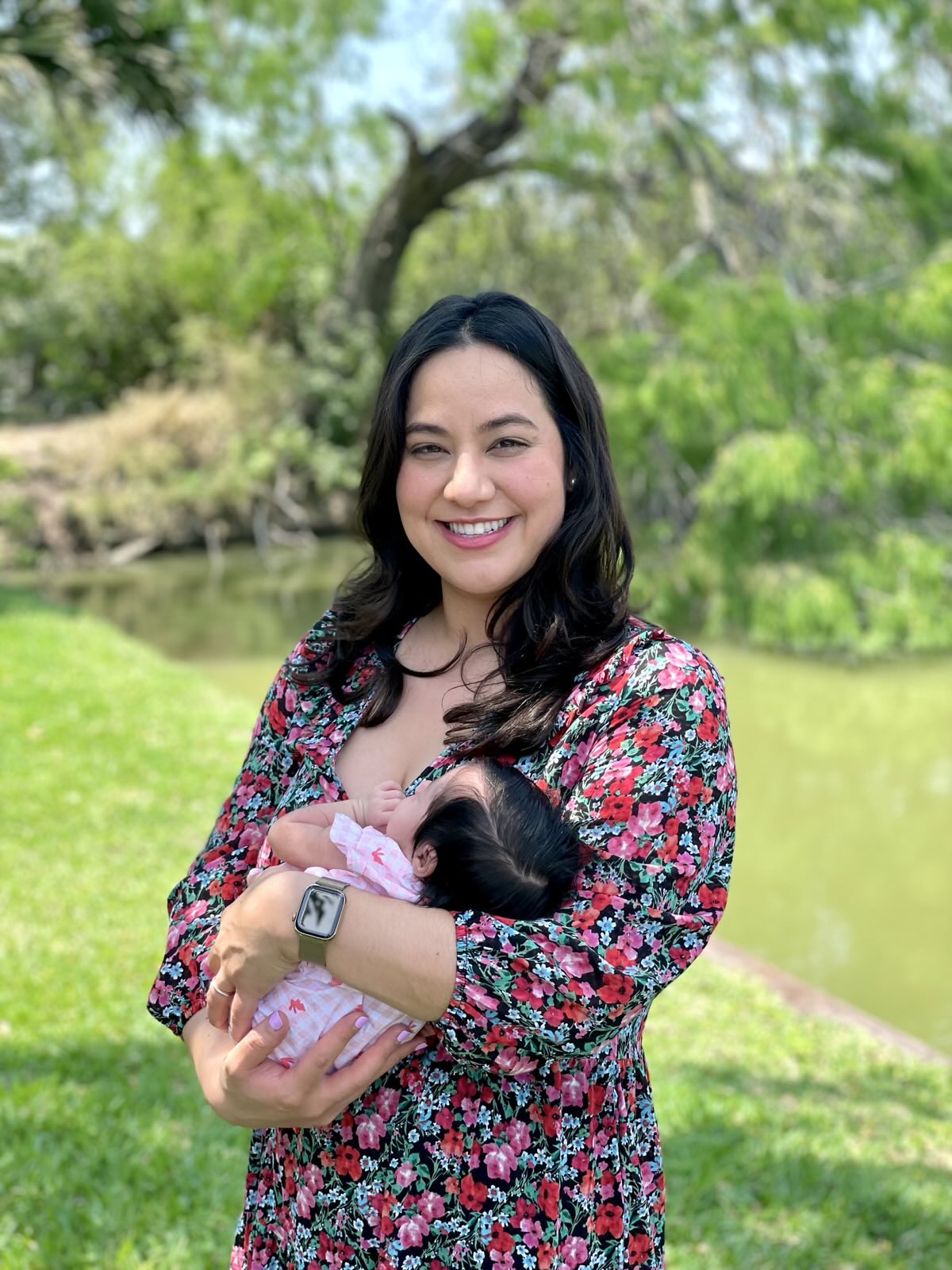 Rochelle Garza stands outdoors in front of a river and trees, smiling as she holds her baby in her arms. She wears a red floral dress and a smartwatch, while the baby is dressed in pink.