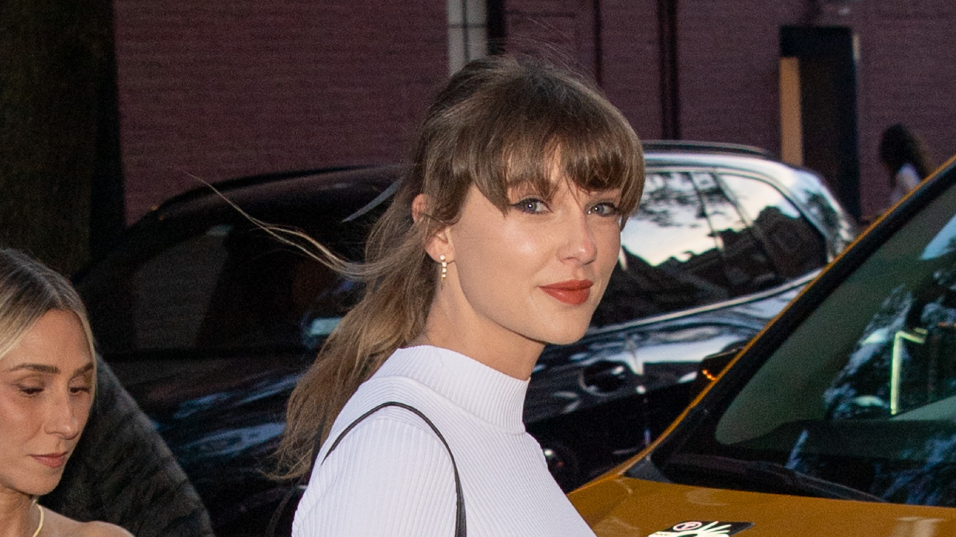 Taylor Swift wearing a white mini dress and black slingback heels and carrying a black low-slung leather bag in New York, standing in front of a yellow cab