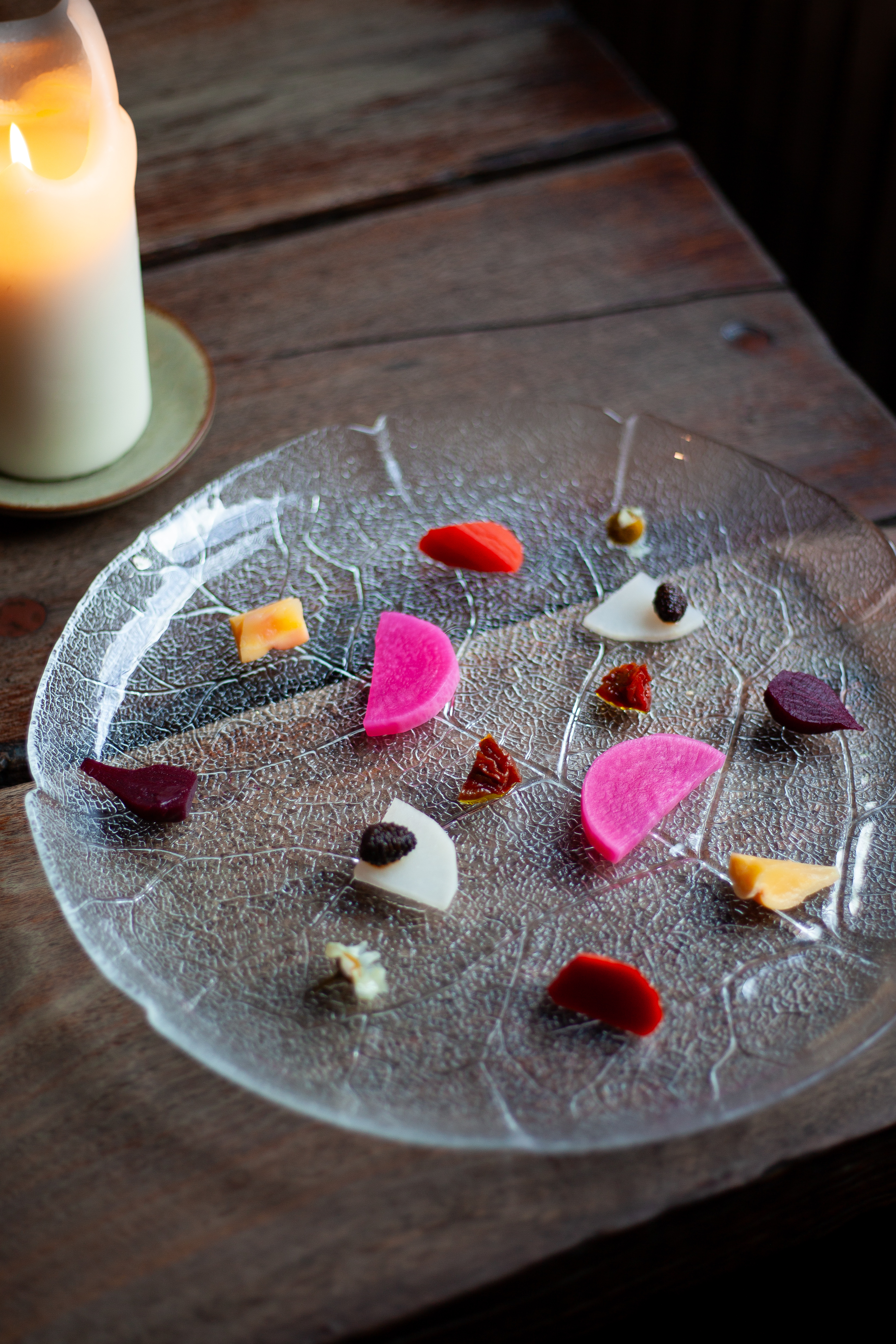 A transparent, leaf-like plate topped with colorful pickled vegetables, placed atop a wooden table and opposite a lit candle.