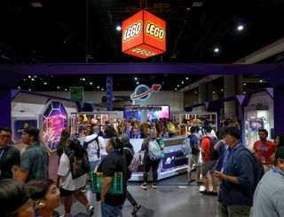 a large group of people congregate on a show floor at a comics convention. a red cube that says "lego" hangs from the ceiling