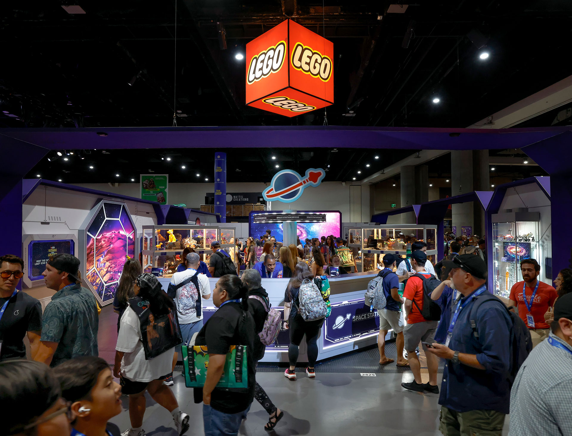 a large group of people congregate on a show floor at a comics convention. a red cube that says "lego" hangs from the ceiling