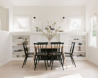 All-white dining room with rectangular, light wood dining table, black wooden dining chairs, low, built-in shelving unit decorated with ornaments and objects beside table, lots of windows creating a light and bright space