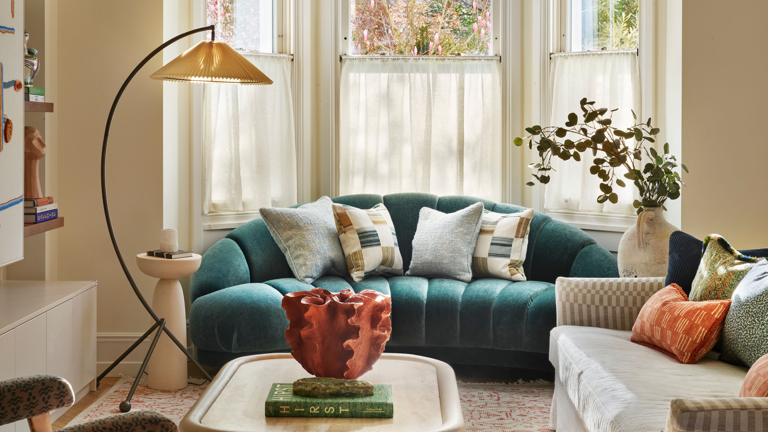 A cozy living room with warm white walls and a bay window with cafe curtains, a teal low-slung sofa in front of the window, a neutral striped sofa with a sculptural coffee table, and patterned rug