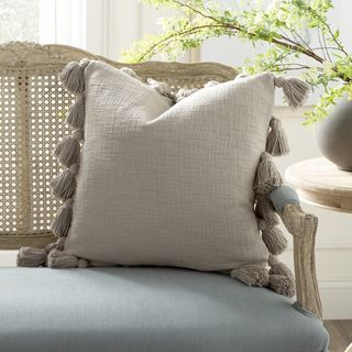 grey square pillow with tassels around the edges sitting on a rattan sofa