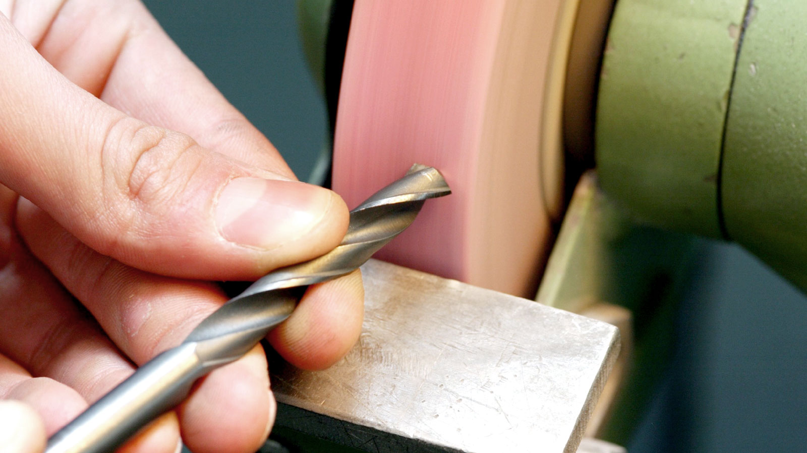 Close up of hand holding twist drill bit up to wheel on bench grinder