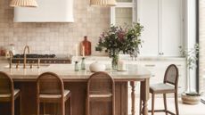 A neutral kitchen with a large wooden and marble island that has four wooden bar chairs around it. The backsplash is made of pink-beige tile and there is white cabinetry and a white hutch