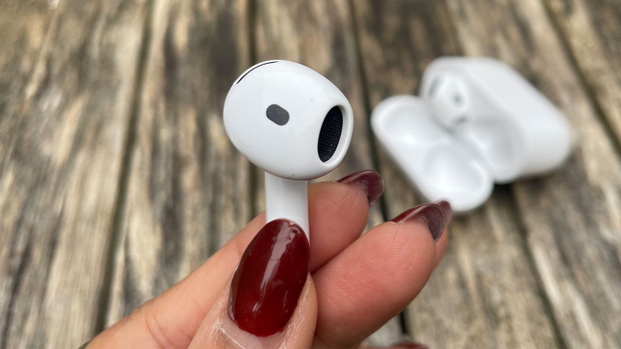 One earbud from the Apple AirPods 4 with ANC held between two fingers and a thumb. In the background, the case is visible with the other earbud inside.