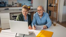 An older couple look happy as they work on paperwork at their dining room table.