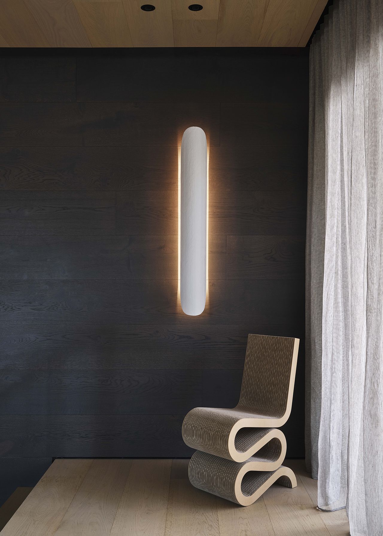 A sculptural chair by a window sits beneath a long, plaster-like sconce