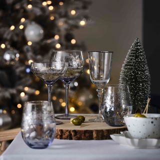 glassware with elegant christmas patterns on them, sitting on top of a wooden tree serving tray, on a table with a white tablecloth