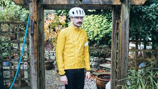 A white man wears a yellow Rapha Core rain jacket waterproof cycling jacket under a trellis