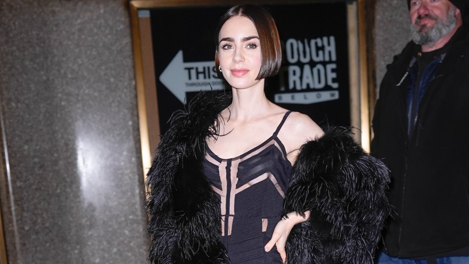 a paparazzi photo of a white woman with a short, dark bob. she is wearing a black dress with cutouts and a black feather coat