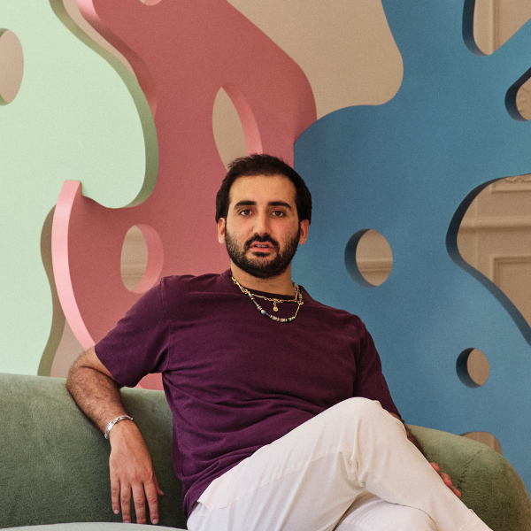 a headshot of Julien Sebban of Uchronia, sitting on a green curved sofa with an interlocking, colorful and abstract scultpure behind, wearing a burgundy-colored t-shirt, white trousers and a necklace with dark short hair and a short dark beard