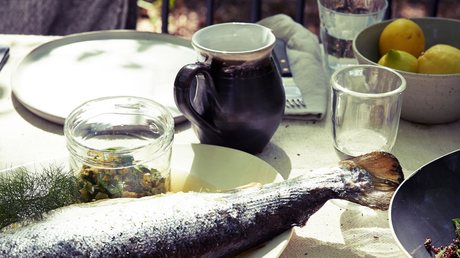 Cooked fish on a table outdoors with a pot of relish, a jug, glass and bowl of lemons