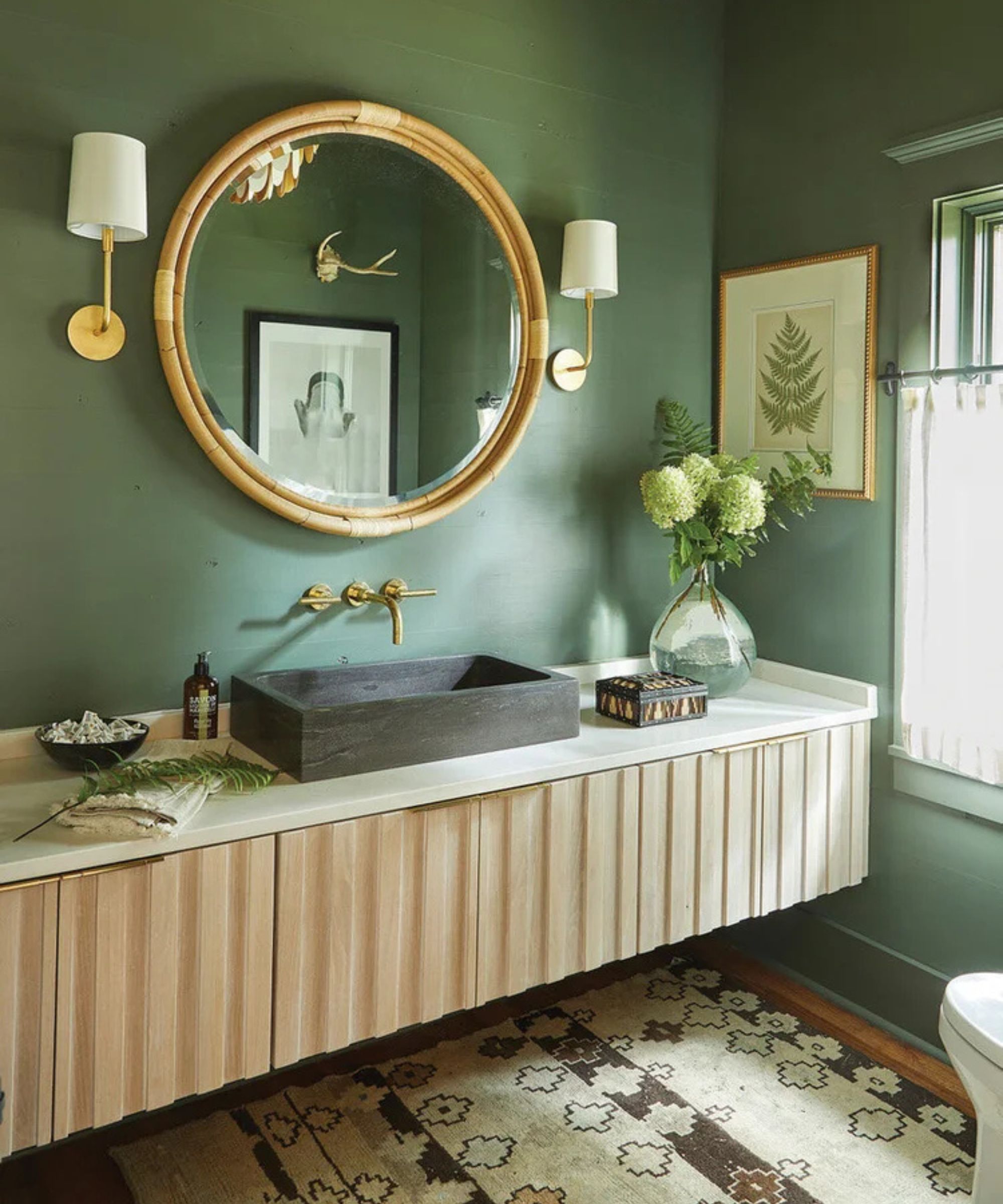 A green bathroom with a floating wooden vanity, a dark concrete sink, and brass accents