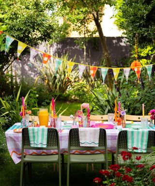 A backyard space with a rectangular table with pink gingham cloth, glasses, colorful candles and floral placemats on it, green and wooden chairs around it, multicolored gingham bunting above it, and trees and fencing