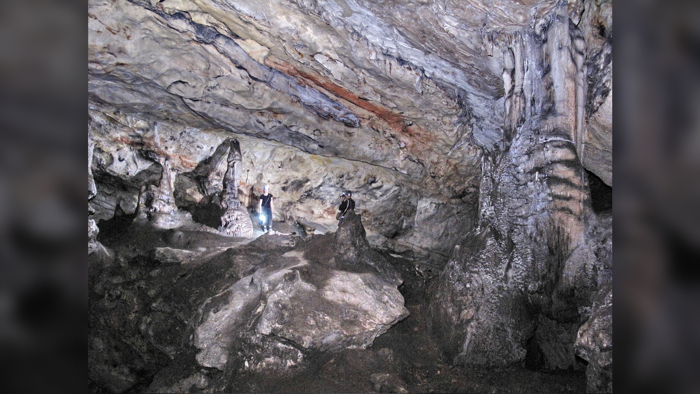 A gray, white and black walls of a cave with a rocky floor and lit with a lamp.