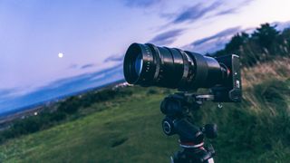 Camera deals: Image shows camera with lens attached,. It's dusk and a field and darkening sky are visible behind the camera.