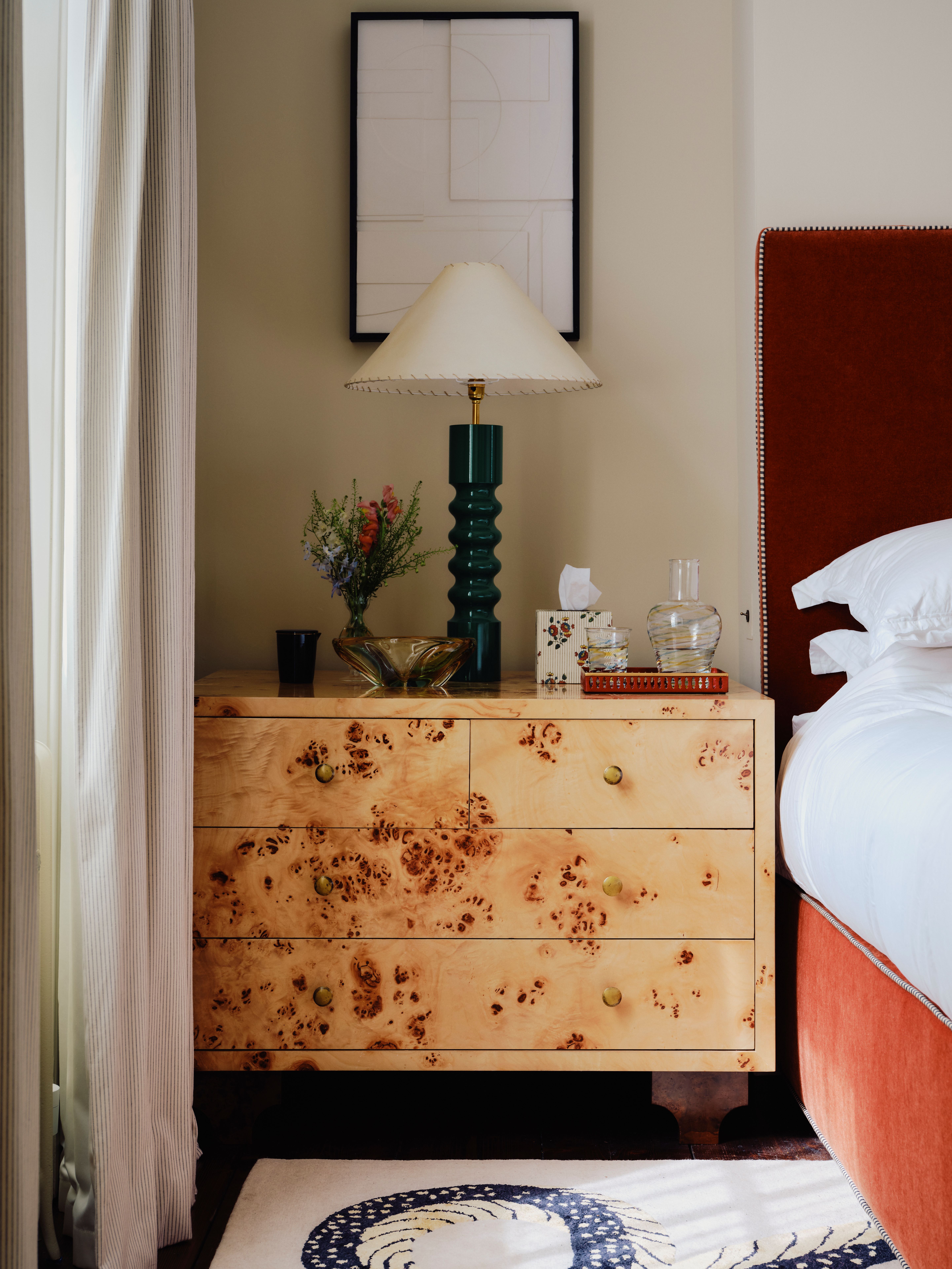 a bedroom with a burl side table with drawers, styled with lamp, dish, tray with carafe and glass, tissue bow, and vase of flowers, with artwork above, and red velvet headboard