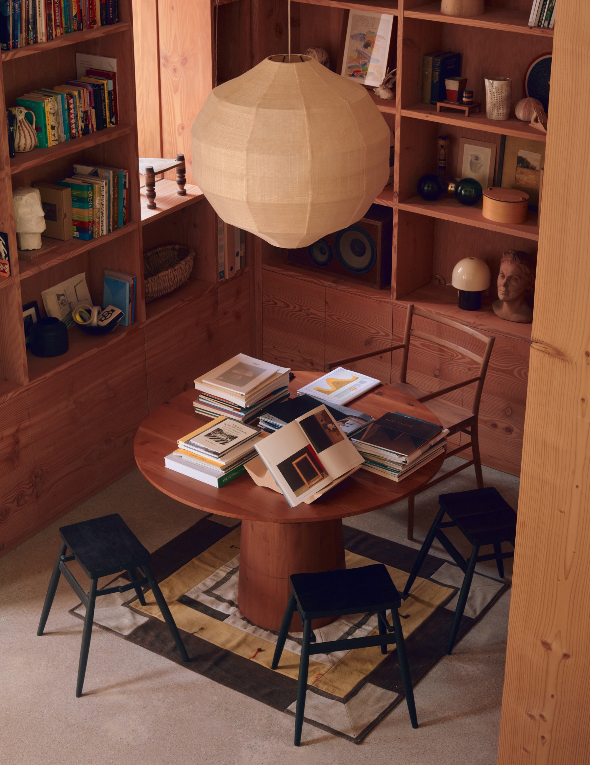 Wood-clad library with built-in shelving, round wood table on top of a rug in the middle with wooden chair and black stools, and paper pendant overhead
