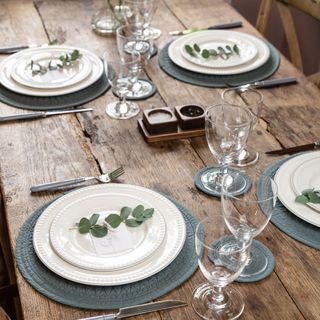 a table set with white plates, glasses and springs of eucalyptus