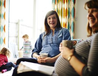 Two heavily pregnant women in lounge enjoying hot drinks and smiling while children play together in background.