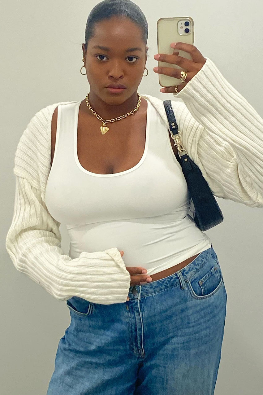 A woman wearing a white tank top, white knit bolero, and blue jeans