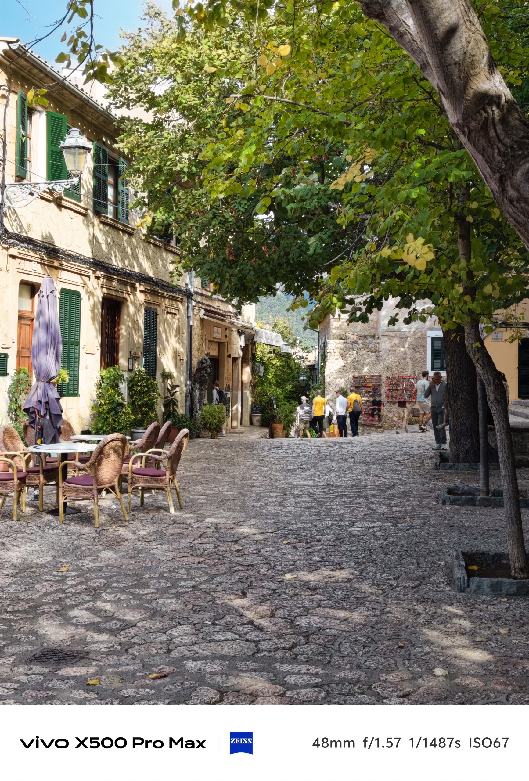 Shaded cobbled village street with cafe tables, stone buildings and leafy trees.