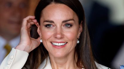 Kate Middleton at a royal event smiling and tucking her hair behind her ears, wearing pearl earrings